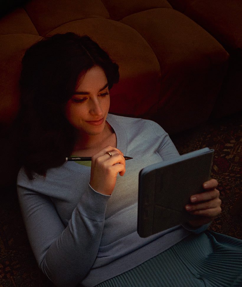 Una donna sdraiata su un divano, illuminata dalla luce soffusa dell’eReader Kobo Libra Colour che tiene in mano, insieme a una Kobo Stylus 2.