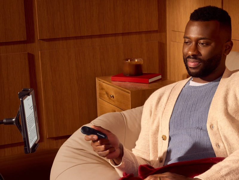 Un uomo, comodamente seduto, tiene in mano il Kobo Remote con la destra, mentre osserva un eReader Kobo su un supporto pieghevole.