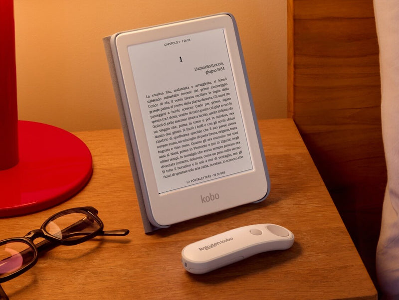 Un Kobo Clara Colour bianco è appoggiato su un tavolo nella sua SleepCover con supporto integrato, accanto a un Kobo Remote bianco e un paio di occhiali.