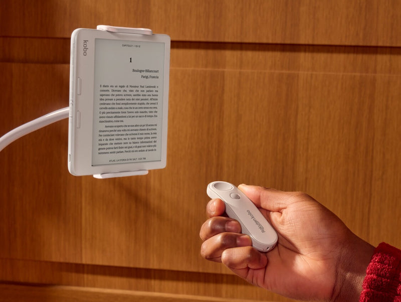 Nella mano destra di una persona è visibile un Kobo Remote bianco, puntato verso un eReader Kobo Libra Colour bianco su un supporto pieghevole.