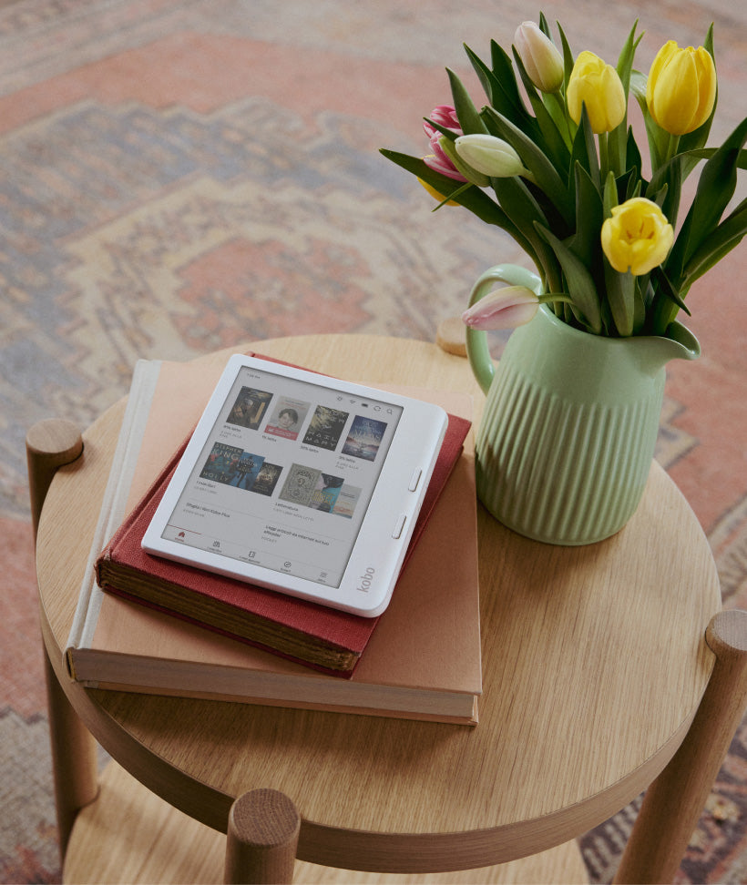 Un eReader Kobo Libra Colour appoggiato su due libri con copertina rigida, sopra un tavolo in legno con un mazzo di fiori in un vaso verde.